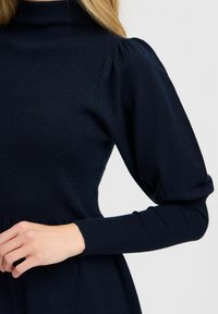 Marinblå polotröja klänning med ribbad textur, puffärmar och figursydd midja. Tyget verkar mjukt och stretchigt. Minimalistisk design.