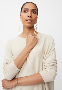 Beige Strickpullover mit übergroßen Ärmeln. Hat einen runden Halsausschnitt und eine weiche Textur. Goldene, runde Ohrringe hängen unter dem Gesicht des Models.