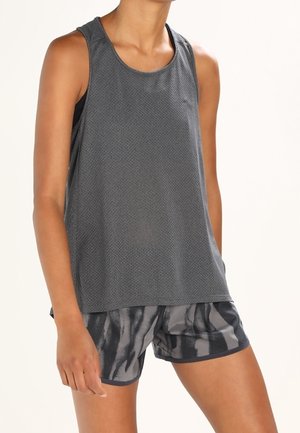Femme portant un haut de sport ample sans manches gris foncé et un short de sport à motif noir et gris sur un fond uni.