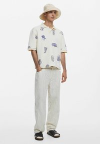 Fehér póló lila mintákkal, világos csíkos nadrággal párosítva. A modellen bézs vödörsapka és fekete szandál van.