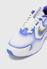 Scarpa atletica bianca e blu con tomaia in mesh, audaci dettagli gialli e suola ammortizzata con design della punta arrotondata.
