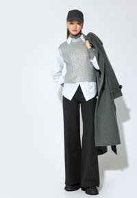 Grijze gebreide vest over een witte blouse, gecombineerd met zwarte broek met krijtstrepen en wijde pijpen. Een grijze pet en jas maken de outfit af. Zwarte schoenen zijn zichtbaar.