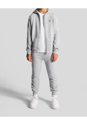 Grijze hoodie met ritssluiting, een voorvak, een wit T-shirt eronder, bijpassende grijze joggingbroek en witte sneakers. De hoodie heeft een gele logo.