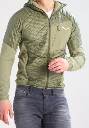 Mand iført olivengrøn quiltet jakke med hætte og grå jeans, justerer ærmet med højre hånd.