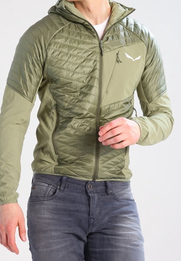 Homme portant une veste matelassée olive à capuche fermée par une fermeture éclair et un jean gris, ajustant la manche avec la main droite.