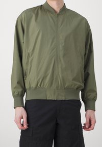 Giacca bomber verde oliva con zip frontale, polsini e orlo a coste, e due tasche laterali; realizzata in un tessuto liscio e leggero.