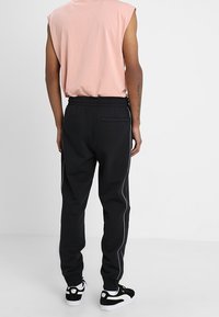 Schwarze Jogginghosen mit elastischem Bund, seitlichem Paspelierdetail und Gesäßtasche. Tragen mit einem pinkfarbenen ärmellosen Shirt und schwarzen Sneakers.