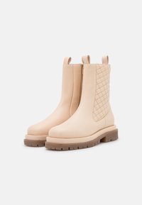 Botas de cuero beige hasta el tobillo con una textura acolchada en un lado, que cuentan con una cremallera lateral y suela de goma gruesa. Acabado suave y tacón plano.