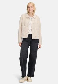 Beige, gestructureerde jas met een kraag, frontknopen en twee grote zakken, gecombineerd met een donkere jeans en enkelboots.