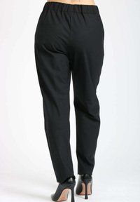 Donna che indossa pantaloni neri con elastico in vita e scarpe con tacco alto nere, vista da dietro su uno sfondo bianco.