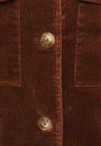 Giacca in cordura marrone con texture rigata, dotata di due tasche frontali e grandi chiusure a bottone. I bottoni sono rotondi con una finitura chiara.