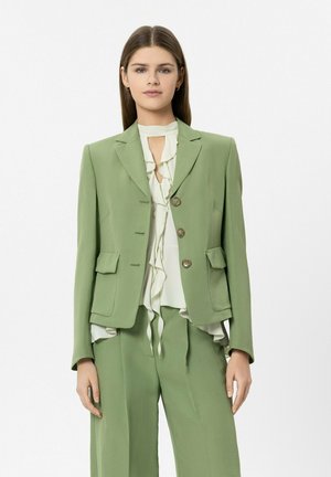 Frau steht in einem hellgrünen, maßgeschneiderten Blazer und passenden Hosen über einer weißen, gerafften Bluse vor einfachem Hintergrund.