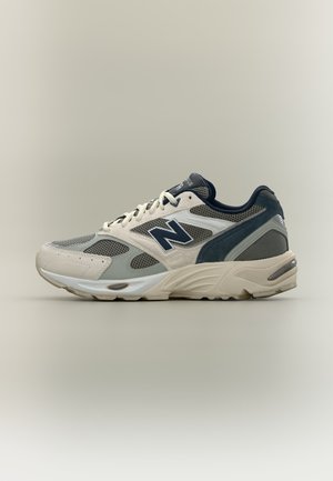 Μπεζ και γκρι αθλητικό παπούτσι New Balance με διχτυωτές επιφάνειες, σκούρο μπλε λογότυπο και λευκά κορδόνια σε ουδέτερο φόντο.