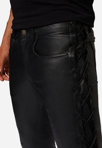 Schwarze Lederhosen mit flachem Frontdesign, seitlichem Schnürdetails und sichtbaren Nähten. Enthält Taschen und einen Metallknopfverschluss.