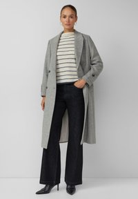 Grauer Wollmantel mit Reverskragen und Knopfverschluss, getragen über einem cremefarbenen Pullover mit navyblauen Streifen, kombiniert mit dunklen, weit geschnittenen Jeans und schwarzen High Heels.