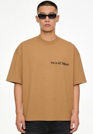 Homme portant des lunettes de soleil noires et un t-shirt marron ample avec le texte "YOU & ME TONIGHT" sur la poitrine sur fond blanc.