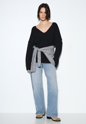 Maglione nero oversize con scollo a V profondo, caratterizzato da una cintura grigia annodata in vita, abbinato a jeans azzurri svasati e stivaletti beige alla caviglia.