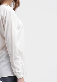 Hvid satinbluse med lange ærmer, glat tekstur og et enkelt design. Har en klassisk krave og afrundet kant.