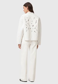 Giacca trapuntata bianca con ritagli a forma di foglia sulla schiena, abbinata a pantaloni bianchi a gamba larga. Texture liscia con una silhouette quadrata.