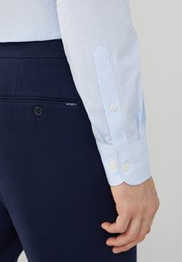 Chemise à carreaux bleus et blancs avec des boutons aux poignets, associée à un pantalon bleu marine foncé avec un bouton et une étiquette logo.
