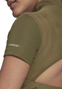 Närbild av en person som bär en olivgrön T-shirt med kort ärm och nätdetalj, med "AEROREADY" tryckt på ärmen.