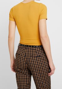 Senapsgul ribbad kortärmad topp i kombination med svart och orange rutig byxa med bälte i midjan och två bakfickor.