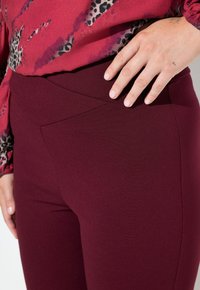 Bordowe legginsy o gładkiej, elastycznej teksturze. Posiadają przekrzywiony pas w talii oraz boczną kieszeń. Zbliżenie z ręką spoczywającą na biodrze.