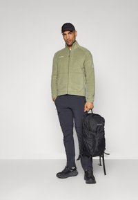 Giacca di pile verde oliva con colletto alto, pantaloni neri con zip e sneakers nere, che tiene uno zaino nero con dettagli del logo.