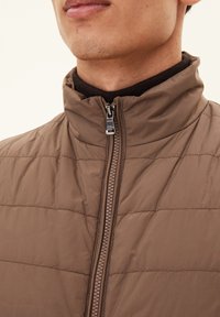 Braune gepolsterte Jacke mit hohem Kragen und Reißverschluss. Sie hat eine gesteppte Textur, eine glatte Vorderseite und eine kontrastierende dunkle Basisschicht.