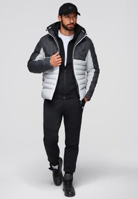 Herrarnas vadderade jacka i svart och ljusgrått med kviltad textur, dragkedjestängning och fast huva, bärs över en svart zip-up tröja och byxor.