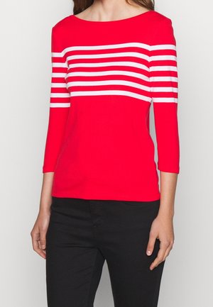 Frau trägt ein rotes Langarmshirt mit weißen horizontalen Streifen auf Brust und Armen, kombiniert mit schwarzer Hose.