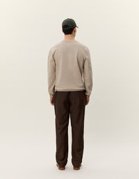 Camisa de manga larga color beige claro combinada con pantalones marrones, vista desde atrás. Una gorra verde oscuro completa el conjunto. Diseño y texturas simples.