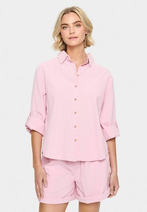 Camisa de botón rosa claro con collar y mangas remangadas, combinada con pantalones cortos a juego. Textura suave, diseño sencillo y botones redondos.