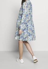 Robe à motifs floraux dans des tons de bleu, vert et marron, avec une jupe à volants à étages, des manches longues bouffantes et une coupe décontractée. Associée à des baskets blanches.