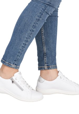 Jambes en jean bleu portant des baskets blanches avec une fermeture éclair latérale et un onglet argenté au talon, debout sur un fond blanc.