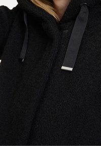 Abrigo negro con capucha hecho de tejido texturizado, que cuenta con cordones negros y un acento metálico. La vista en primer plano muestra el material y la construcción.