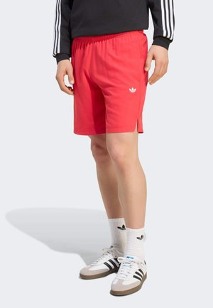 Persona che indossa pantaloncini rossi Adidas, calzini bianchi con logo nero, scarpe da ginnastica Adidas nere e bianche e una maglia nera a maniche lunghe con strisce bianche.