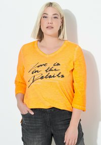 Donna con capelli biondi che indossa una maglietta arancione con la scritta "L'amore è nei dettagli" e jeans neri, in piedi contro un muro bianco.