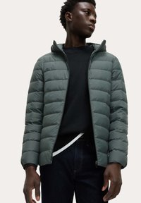 Groene puffer jacket met horizontaal gestikte secties, ritssluiting en een capuchon. Draagt over een zwart shirt en blauwe jeans. Minimalistisch ontwerp.