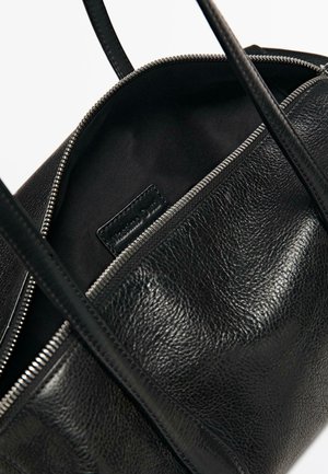 Borsa a mano in pelle nera con cerniera argentata parzialmente aperta, che mostra l'interno in tessuto nero e l'etichetta del marchio all'interno.