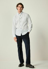 Camisa blanca de botones con pequeños lunares azules, de mangas largas y un bolsillo, combinada con pantalones oscuros navy y zapatos de vestir negros.