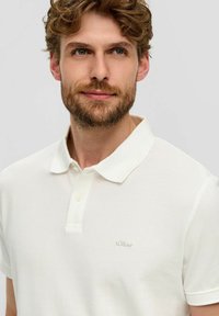 Mann mit lockigem braunem Haar und Bart, der ein weißes Poloshirt mit kurzen Ärmeln vor einem schlichten hellen Hintergrund trägt.