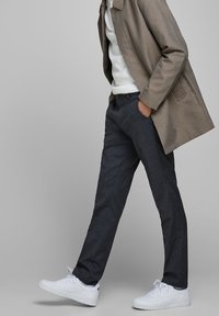 Grå rutig kappa över en vit stickad tröja, i kombination med mörka skräddarsydda byxor och vita sneakers. Slät tyg, strukturerad passform och modern design.