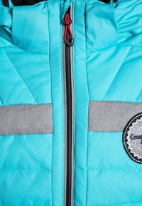 Veste matelassée turquoise avec des rayures horizontales grises, fermeture éclair et patch logo rond, avec un tissu texturé et une capuche réglable.