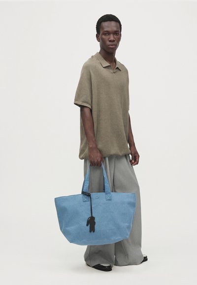 Un homme porte un polo couleur taupe et un pantalon gris. Il tient un grand sac cabas bleu orné d’un pendentif noir, sur un fond de studio uni.