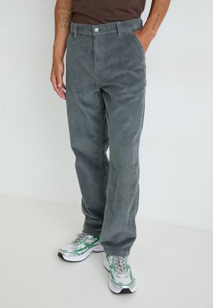 Osoba nosząca szare sztruksowe spodnie i białe trampki z zielonymi akcentami, stojąca z rękami w kieszeniach na białym tle.