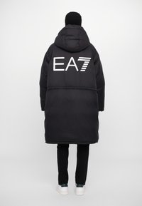 Melnā polsterētā garā mētli ar kapuci, uz kuras atrodas liels balts EA7 logo uz aizmugures. Gluda tekstūra, elastīgas aproces un taisna piegriezuma.
