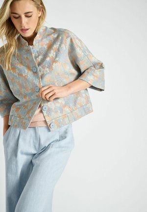 Veste à manches courtes en tissu bleu clair avec un motif floral en pêche et rouille. Cinq boutons bleus et un col rond.