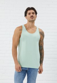 Mátově zelený tílko s kulatým výstřihem, volný střih a měkká bavlněná tkanina. Modelka má na sobě světle modré džíny, které odhalují tetování na paži.
