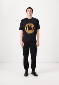 Sort kortærmet t-shirt med et gult grafisk design, iført sorte joggerbukser og sorte sko, mod en ensfarvet hvid baggrund.